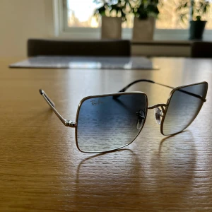 Ray-Ban 1971 Square solglasögon - Snygga Ray-Ban 1971 Square solglasögon med fyrkantiga linser och en tunn silverfärgad metallram. Glasen har en gradient som går från mörkare till ljusare ton. Perfekta för en stilren look. Säljer pga felköp förra sommaren, de är aldrig använda. Nypris ca 1700