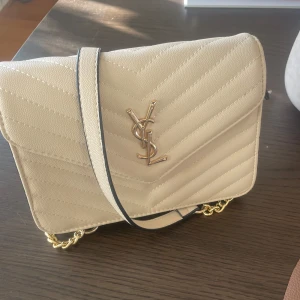 Beige axelväska från YSL - Snygg beige axelväska från YSL med quiltad design och guldfärgade detaljer. Väskan har en elegant kedja och en ikonisk logotyp på framsidan. Perfekt för att ge en lyxig touch till din outfit.