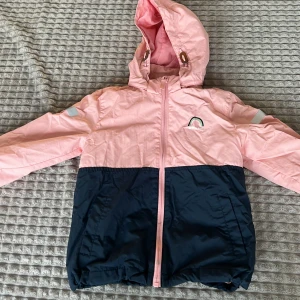 Rosa och marinblå jacka med huva - Säljer en söt jacka i rosa och marinblått med huva och dragkedja. Jackan har långa ärmar och en liten regnbågsdetalj på bröstet. Perfekt för vår och höst!