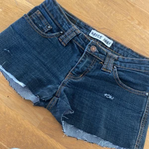Mörkblå jeansshorts från Miss Anna - Snygga mörkblå jeansshorts från Miss Anna med fransiga kanter och slitna detaljer. Perfekta för en avslappnad stil. Klassisk femficksdesign och knappgylf.