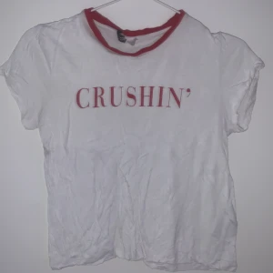 Vit t-shirt med röd text 'CRUSHIN'' - Säljer en vit t-shirt från H&M med röd text 'CRUSHIN'' på framsidan. T-shirten har en röd kant runt halsen och är kortärmad. Perfekt för en avslappnad stil!