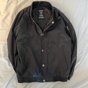 Svart bomberjacka från Someday Soon - Riktigt tuff och snygg svart bomberjacka från Someday Soon med texten 'I WILL CHANGE THE WORLD' på ryggen. Jackan har dragkedja och knappar framtill samt ribbade muddar. Tillverkad i 100% polyester, perfekt för en cool och stilren look. Medela vid minsta funderingar! 👍