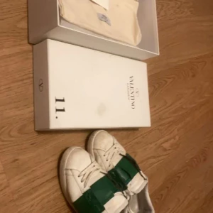 Valentino Garavani sneakers med grönt band - Säljer ett par stilrena Valentino Garavani sneakers i vitt med ett iögonfallande grönt band över snörningen. Skorna kommer med originalförpackning och dustbag. Perfekta för att ge din outfit en lyxig touch.
