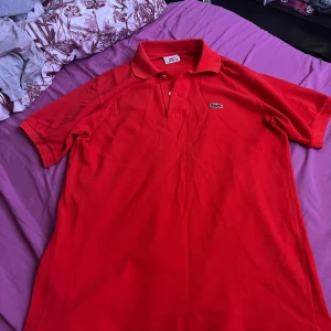 orange pikétröja från Lacoste - Säljer en klassisk röd pikétröja från Lacoste med korta ärmar och den ikoniska krokodilloggan på bröstet. Perfekt för en stilren och sportig look. Tröjan har en krage med knappar för en justerbar passform.