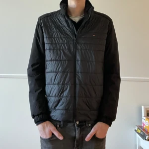 Svart hybrid jacka från Tommy Hilfiger - Snygg svart hybrid jacka från Tommy Hilfiger med dragkedja framtill. Jackan har långa ärmar och en stilren design, perfekt för mellansäsongen. Den har en diskret logga på bröstet och ribbade muddar vid ärmslut.