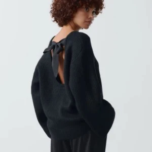 Svart stickad tröja med öppen rygg och rosett - Snygg svart stickad tröja med en elegant öppen rygg och en dekorativ rosett. Perfekt för att ge en stilfull touch till din outfit. Tröjan har långa ärmar och en bekväm passform.