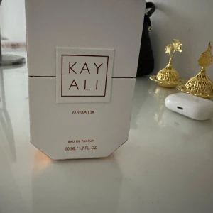 Kayali Vanilla 28 Eau de Parfum - Kayali Vanilla 28 är en lyxig Eau de Parfum med en varm och söt doft av vanilj. Flaskan är elegant med en djup bärnstensfärg och en kristallklar kork. Förpackningen är stilren i vitt med guldiga detaljer. Perfekt för den som älskar en sofistikerad och inbjudande doft.