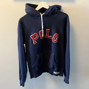 Mörkblå hoodie från Ralph Lauren - Säljer en snygg mörkblå hoodie från Ralph Lauren med texten 'POLO' i rött och vitt på framsidan. Tröjan är sällsynt och har en klassisk känguruficka och justerbar huva med vita snören. Den är sliten vid handlederna men annars är den bra. Perfekt för en avslappnad stil.