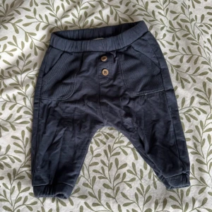 Mörkblå byxor från Newbie - Säljer ett par mörkblå byxor från Newbie i mjukt material. Byxorna har elastisk midja och muddar vid bensluten för extra komfort. Två knappar framtill och en liten ficka bak ger en söt detalj.