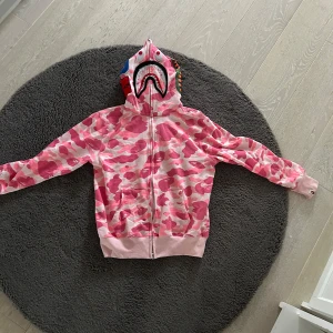 Rosa BAPE tröja - Säljer en rosa BAPE camo hoodie med dragkedja och unikt mönster på huvan. Tröjan har långa ärmar och ribbade muddar. Perfekt för en cool och avslappnad stil. 
