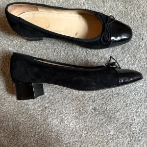 Svarta ballerinaskor med klack - Eleganta svarta ballerinaskor med låg klack och spetsig tå. Skorna har en kombination av mocka och skinn, med en dekorativ rosett framtill. Perfekta för en stilren look.