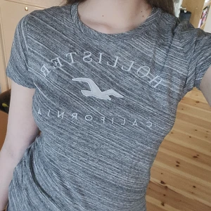 Grå t-shirt från Hollister - Säljer en gråmelerad t-shirt från Hollister med tryck framtill. T-shirten har en normal passform och korta ärmar, perfekt för en avslappnad stil. Skön och enkel att matcha med jeans eller shorts.