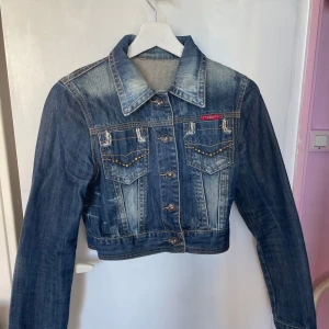 Blå croppad jeansjacka - Säljer en snygg blå croppad jeansjacka från Levi's. Jackan har långa ärmar och knappar framtill. Den är dekorerad med nitar på fickorna och har en klassisk krage. Perfekt för en trendig look!
