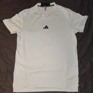 Vit kortärmad t-shirt från Adidas - Säljer en stilren vit t-shirt från Adidas med deras klassiska logga på bröstet. Perfekt för träning eller en avslappnad dag. T-shirten är kortärmad och gjord i ett bekvämt material.
