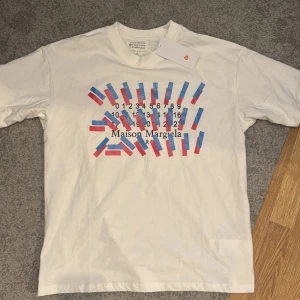 Vit t-shirt från Maison Margiela - Säljer en tvär fet vit t-shirt från Maison Margiela med ett unikt tryck i blått och rött. Storleken är M. Den är i 10/10 skick (helt ny). Säljer för att den var för stor. Skriv vid funderingar!💯