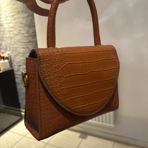 Brun handväska med krokodilmönster - Snygg brun handväska i syntet med krokodilmönster. Väskan har ett elegant handtag och en praktisk magnetknäppning. Insidan är röd och har en dragkedja för extra säkerhet. Perfekt för att ge din outfit en stilfull touch.