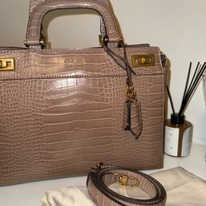 Brun handväska från Guess - Aldrig använt  Elegant handväska från Guess i en vacker, ljusbrun färg med krokodilmönster. Väskan har ett handtag och en avtagbar axelrem. Den har även en dekorativ berlock med ett litet hänglås och en nyckel. Insidan är rymlig med flera fack och dragkedjor för säker förvaring.