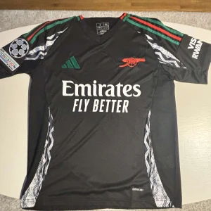 Arsenal Bortaställ 24/25 - Säljer en svart fotbollströja från Adidas med tryck på bröstet och ärmarna. Tröjan har korta ärmar med gröna och röda detaljer samt ett UEFA-märke. Perfekt för fotbollsfans!
