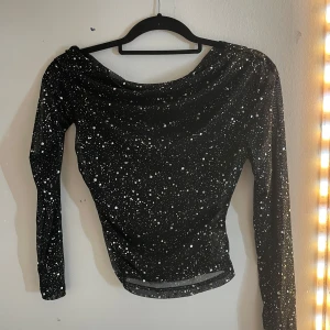 Svart glittrig topp  - Säljer en svart glittrig topp med långa ärmar och ett stjärnliknande mönster. Perfekt för en kväll ute eller festliga tillfällen. Toppen har en elegant passform och är tillverkad i ett syntetmaterial som ger en skön känsla.