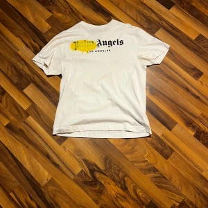 Vit t-shirt från Palm Angels - Säljer en vit t-shirt från Palm Angels med tryck i svart och gult på framsidan. T-shirten har en avslappnad passform och är kortärmad. Perfekt för en casual look.