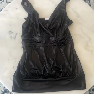 Svart vintage topp❤️ - Snygg svart topp med v-ringning och ärmlös design. Toppen har en elegant glansig finish och rynkade detaljer i midjan för en smickrande passform. Perfekt för en kväll ute eller en speciell tillställning.
