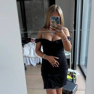 Svart klänning med spetsdetaljer - Snygg svart klänning med detaljer på axelbanden. Klänningen har en tight passform och är perfekt för en kväll ute. Den har en kort längd och volangdetaljer vid fållen. S/M