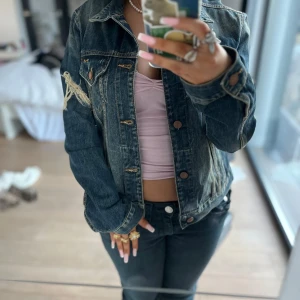 Blå jeansjacka med slitningar - Snygg blå jeansjacka med slitna detaljer och knappar framtill. Jackan har en klassisk design med två bröstfickor och långa ärmar. Perfekt för en avslappnad stil.