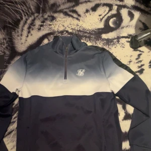 Blå och vit tröja med dragkedja från Siksilk - Snygg blå och vit tröja från Siksilk med halv dragkedja och logga på bröstet. Perfekt för en stilren look. Tröjan har långa ärmar och en gradvis färgövergång från mörkblått till vitt.