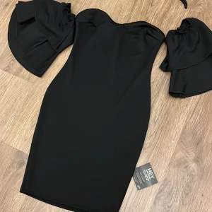 Svart klänning från Nelly - Snygg svart klänning från Nelly med offshoulder-design och volangdetaljer på ärmarna. Perfekt för en elegant kvällslook. Klänningen har en tight passform som framhäver figuren.