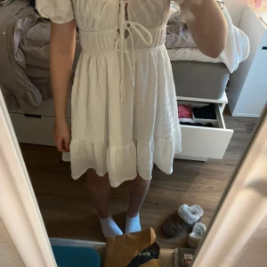 Vit klänning från Gina Tricot - Säljer en söt vit klänning från Gina Tricot med knytning framtill och volangdetaljer. Klänningen har korta ärmar och en luftig passform, perfekt för varma dagar.