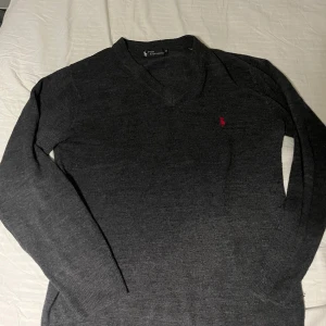 Grå tröja från Polo Ralph Lauren - Säljer en stilren grå tröja från Polo Ralph Lauren med v-ringning och ett diskret rött broderat märke på bröstet. Tröjan har långa ärmar och är perfekt för en klassisk look.