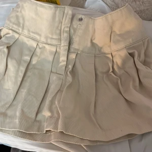 Beige kjol med stjärndetalj - Säljer en beige kjol med en unik stjärndetalj på baksidan. Kjolen har en dragkedja och knapp framtill för enkel påklädning. Den är kort och har en veckad design som ger en fin volym.