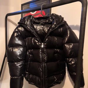 Svart pufferjacka från Zara - Snygg svart pufferjacka från Zara med dragkedja och huva. Perfekt för kyliga dagar med sin quiltade design som håller värmen. Jackan har en glansig finish och är ett stilrent val för vintern.
