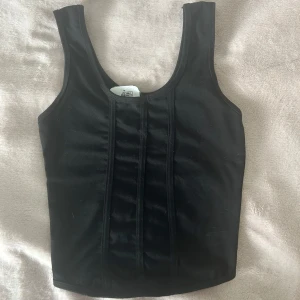 Svart korsettliknande topp - Snygg svart korsettliknande topp med breda axelband och en stilren design. Perfekt för att ge en edgy touch till din outfit. Passar bra till både jeans och kjol.