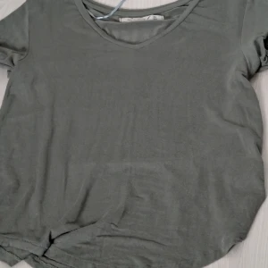 Grå t-shirt med v-ringning - Säljer en stilren grön t-shirt med v-ringning. Perfekt för en avslappnad look. T-shirten har korta ärmar och är tillverkad i ett mjukt material.