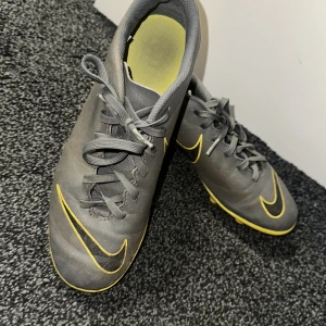 Grå och gula fotbollsskor från Nike - Snygga grå fotbollsskor från Nike med gula detaljer och snörning. Skorna har en slitstark sula med dobbar, perfekt för spel på gräs. Den ikoniska Nike-loggan syns tydligt på sidan och sulan.