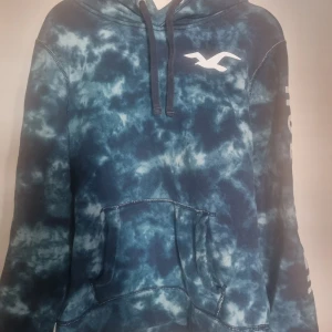 Blå hoodie från Hollister - Blå hoodie från Hollister med batikmönster. Tröjan har en stor vit logga på bröstet och en känguruficka framtill.