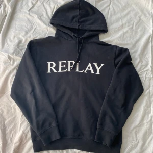 Svart hoodie från Replay - Säljer en svart hoodie från Replay med stor vit logga på framsidan. Den har en justerbar luva med snören och ribbade muddar vid ärmslut och nederkant. Perfekt för en avslappnad stil. Har en defekt vid snörena som syns på bild 3. Vid frågor och funderingar är den bara att skicka ett dm💫