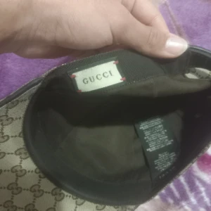 Beige keps från Gucci - Snygg beige keps från Gucci med det klassiska GG-mönstret. Kepsen har en justerbar rem baktill och en grön och röd randdetalj på sidan. Perfekt för att ge en lyxig touch till din outfit.