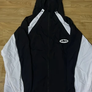 Svart och vit vindjacka från Cortez - Säljer en stilren svart och vit vindjacka från Cortez med huva och dragkedja. Jackan har ett sportigt utseende med vita ärmar och en logotyp på bröstet. Perfekt för blåsiga dagar.