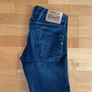 Replay Anbass Mörkblå - Snygga mörkblå jeans från Replay med klassisk femficksdesign och knappgylf. Jeansen har en diskret broderad detalj på bakfickan och är perfekta för en stilren look. Det är slim fit och de är i mycket gott skick.