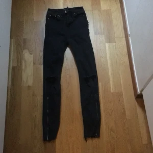 Svarta jeans med dragkedja och slitningar - Snygga svarta jeans med slitningar på knäna och dragkedjor vid anklarna. De har en tight passform och är perfekta för en trendig look. Högmidjade med klassisk femficksdesign. Jeansen är helt oanvända då de är köpte små. Jeansen är mindre i passform
