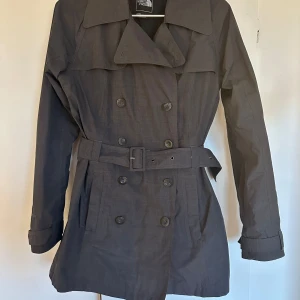 Svart regnjacka/trenchcoat från The North Face - Snygg svart regn trench från The North Face med bälte i midjan och knappar framtill. Perfekt skick, som ny