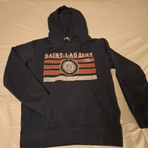 Mörkblå hoodie från Saint Laurent - Säljer en snygg mörkblå hoodie från Saint Laurent med tryck i rött och vitt på framsidan. Hoodien har en klassisk känguruficka och justerbar huva med snören. Perfekt för en avslappnad stil. Strl M. Hör av dig vid frågor. Kan gå ner i pris vid snabb affär!