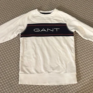 Vit tröja från Gant - Säljer en stilren vit tröja från GANT med ett mörkblått band och röd detalj över bröstet. Skick 10/10 || storlek 146-152/11-12 år. Tröjan har långa ärmar och en rund halsringning. Perfekt för en avslappnad stil. Priset kan diskuteras vid snabb affär.