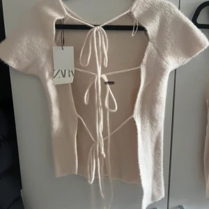 Beige kortärmad topp från Zara - Säljer en superfin beige topp från Zara med korta ärmar och knytning i ryggen. Toppen har en mjuk och fluffig textur som ger en mysig känsla. Perfekt för en stilren look!