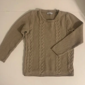 Beige stickad tröja - Säljer en mysig beige stickad tröja från Freja med flätmönster på framsidan. Tröjan har långa ärmar och en rund halsringning. Perfekt för kyliga dagar!