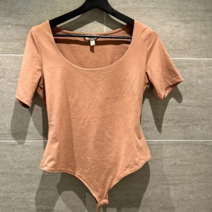 Beige bodysuit från H&M - Säljer en stilren beige bodysuit från H&M med korta ärmar och rund halsringning. Perfekt för att matcha med jeans eller kjol för en enkel och snygg look. Materialet är mjukt och stretchigt för en bekväm passform.