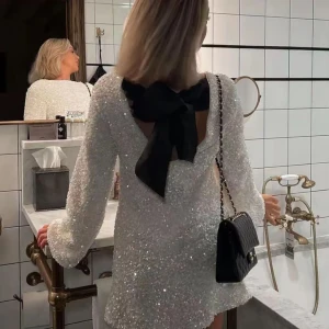 Vit glittrig klänning med öppen rygg och rosett - Snygg vit klänning med glittrande paljetter och en elegant öppen rygg. Klänningen har långa ärmar och en stor svart rosett i nacken som ger en extra touch av glamour. Perfekt för en festlig kväll! Säljer då den inte kommer till användning.