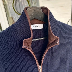 Mörkblå stickad väst från Gran Sasso - Snygg mörkblå stickad väst från Gran Sasso med dragkedja och bruna detaljer. Perfekt för lager-på-lager under kyliga dagar. Den har en stilren design med ribbad struktur och bruna kantdetaljer vid kragen och fickorna.